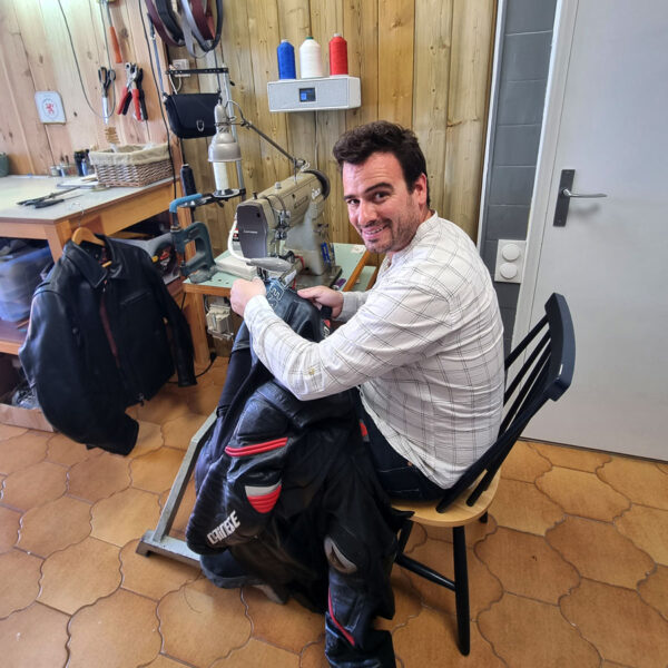 Alexandre Vigouroux, artisan couturier, souriant derrière sa machine à coudre. Veste en cuir pour motocyclistes, atelier chaleureux en bois. Services de retouche et réparation de vêtements en cuir. #couture #artisanat #motocyclisme